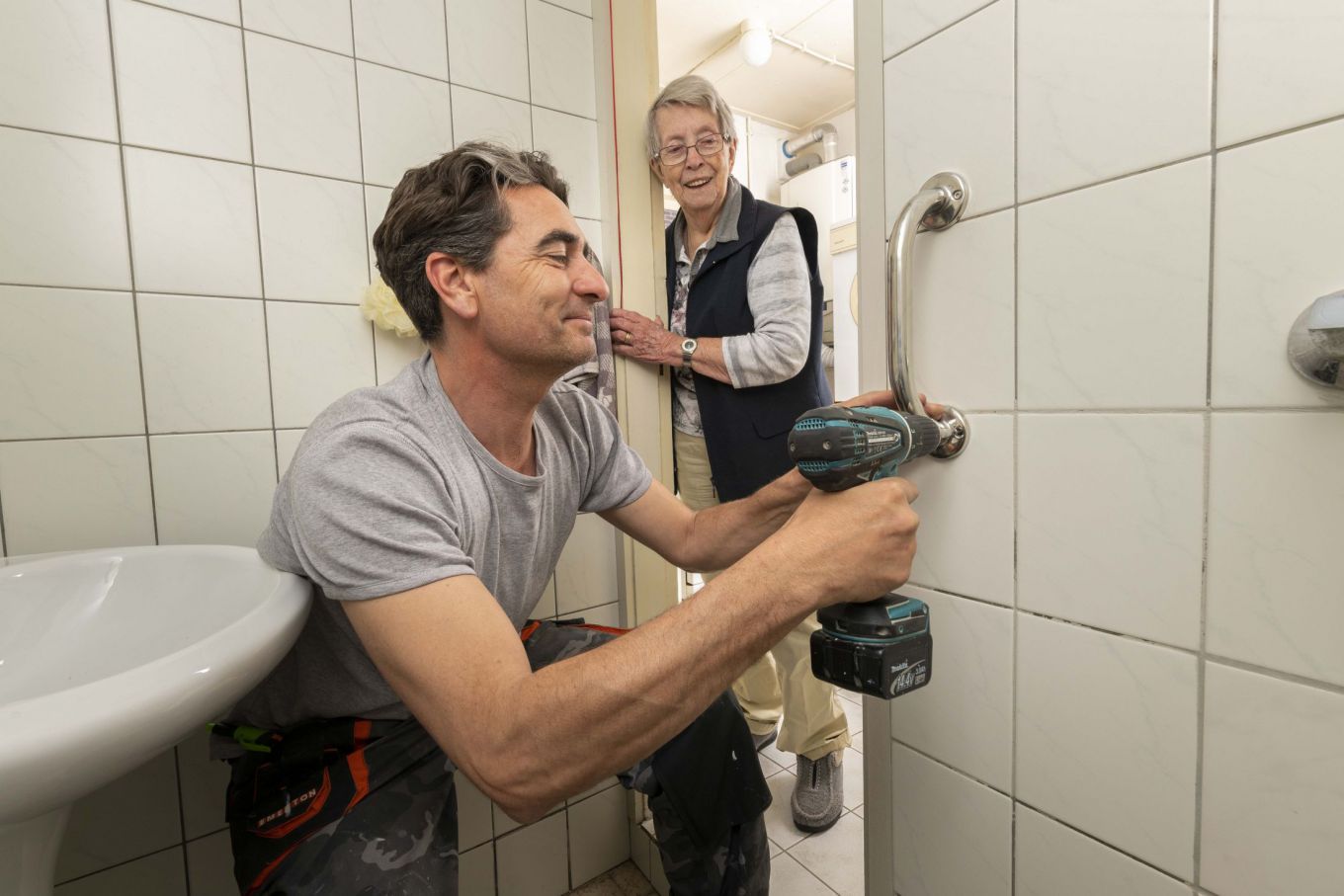 Foto van man die in de badkamer een handgreep in de muur schroeft terwijl een oudere vrouw op de achtergrond blij toekijkt