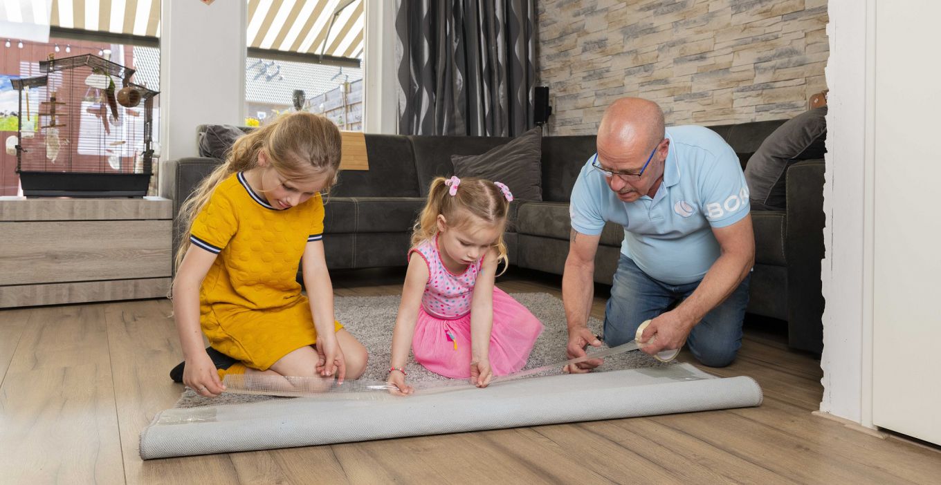Foto van een man en twee kinderen die op de grond zitten en tape op de onderkant van het tapijt bevestigen